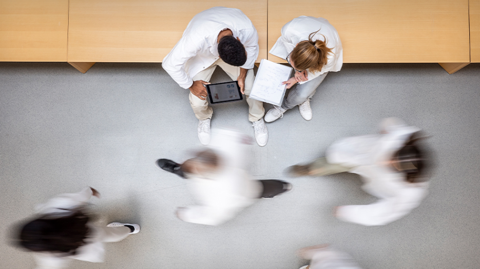 Bird's eye view of 2 clinicians sitting on a bench with blurred figures walking past them.
